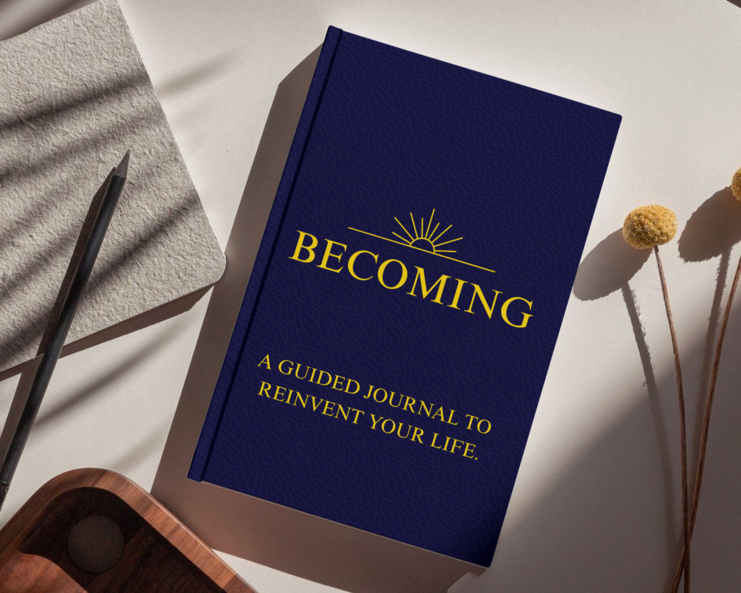 Blue book titled 'Becoming' on a white surface with a pen and dried flowers.